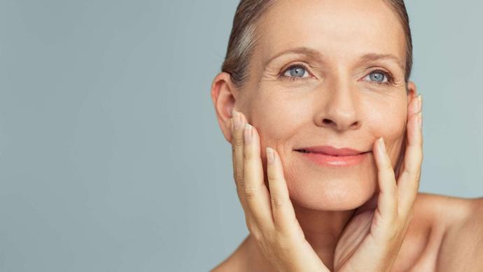 Visage de femme senior avec une belle peau qui sourit et se tient les joues avec les mains, sur fond gris clairit en se tenan