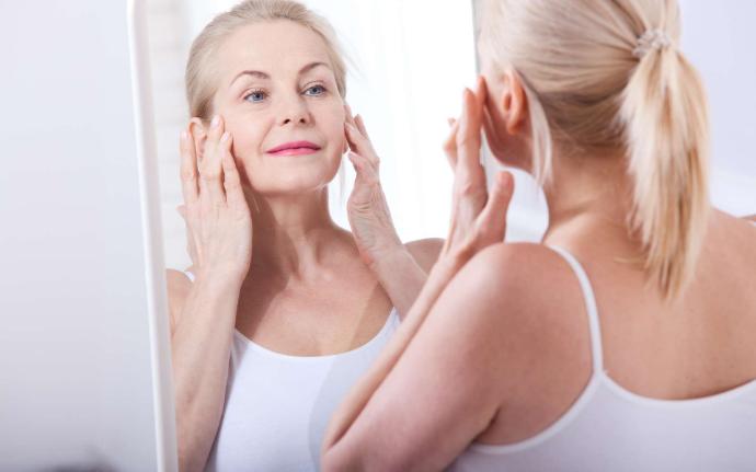 Femme senior blonde avec queue de cheval qui se regarde dans le miroir en touchant ses joues avec ses mains