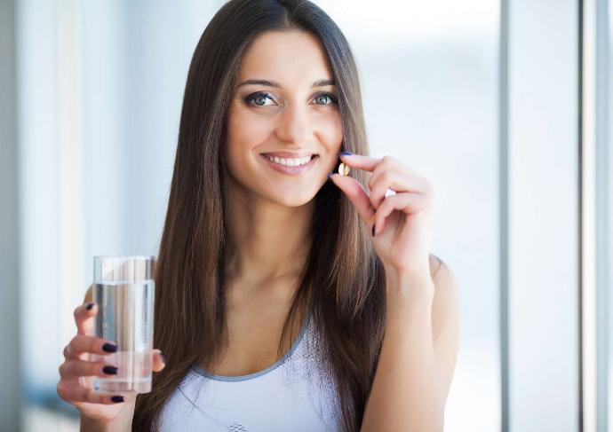 Jeune femme brune qui tient d'une main un verre d'eau et de l'autre un complément alimentaire