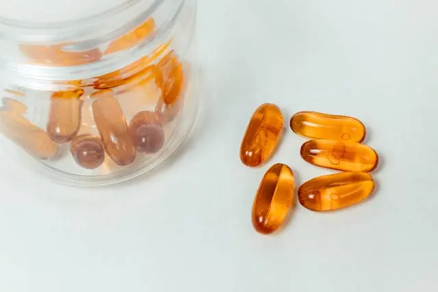 Bocal transparent avec capsules compléments alimentaires oranges et 5 capsules oranges à côié sur fond vert d'eau