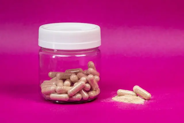 Gélule dans pot transparent avec couvercle blanc avec deux gélules et de la poudre à côté sur fond rose fushia