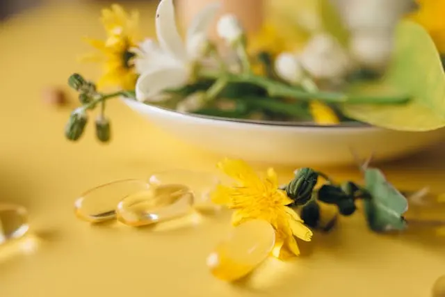 Compléments alimentaires en gélules sur surface jaune avec en arrière-plan une coupelle avec fleurs blanches et jaunes