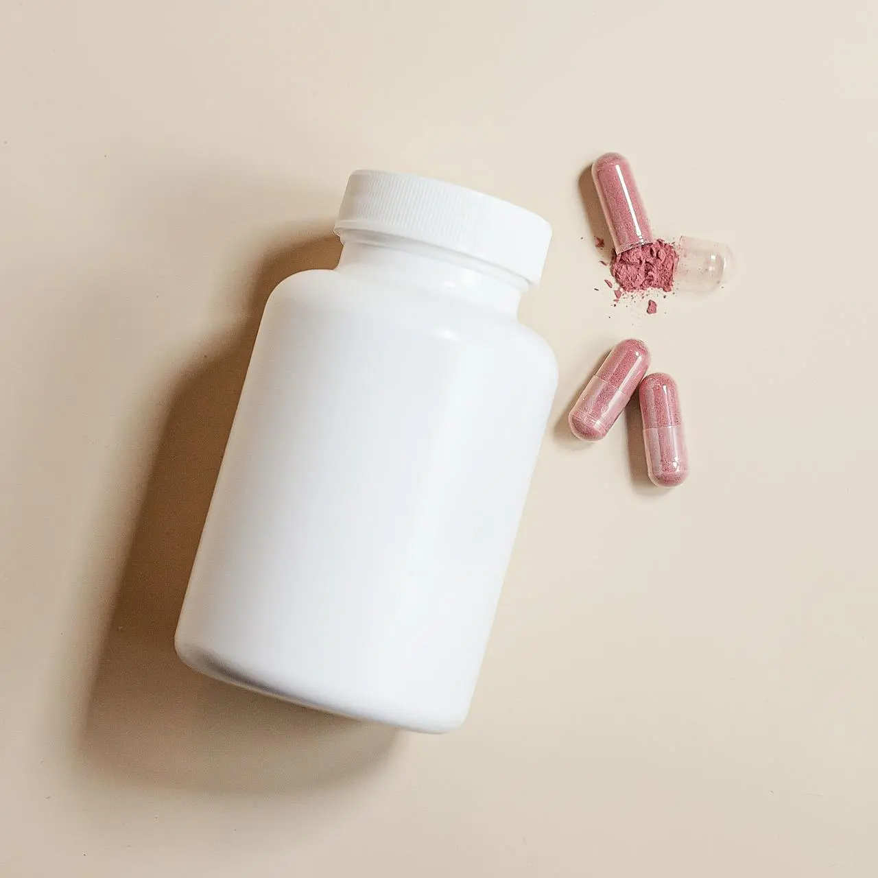 Flacon blanc compléments alimentaires avec trois gélules roses et une ouverte avec poudre rose sur fond beige